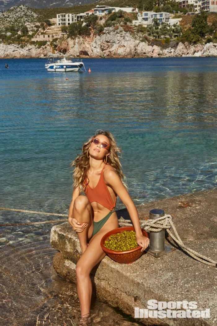 Olivia Ponton sits on a concrete dock on the edge of the water in a rust red and green-colored one-piece.